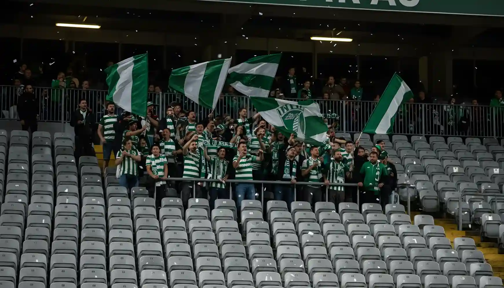 Pequeño grupo de aficionados visitantes en una sección del estadio rival con banderas verdes y blancas